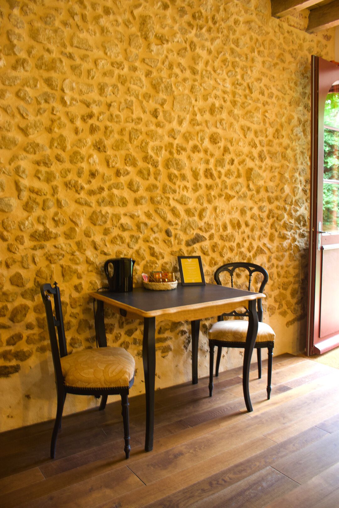 Un muro de piedra, una mesa de madera, dos sillas y una mesita con objetos encima.