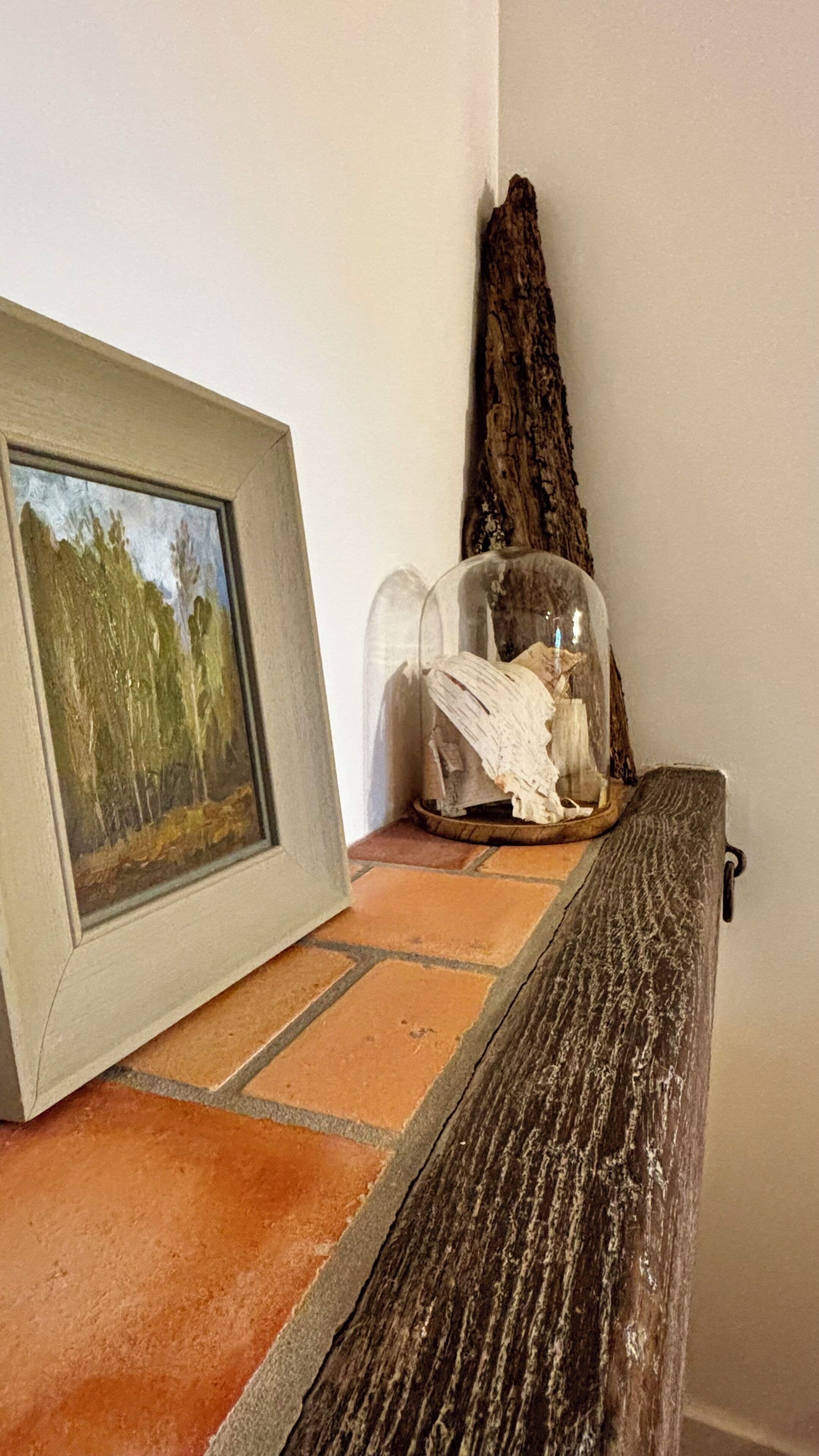Um quadro emoldurado sobre uma superfície revestida de azulejos, um recipiente de vidro transparente com um objeto branco, e um pedaço de madeira encostado na parede.