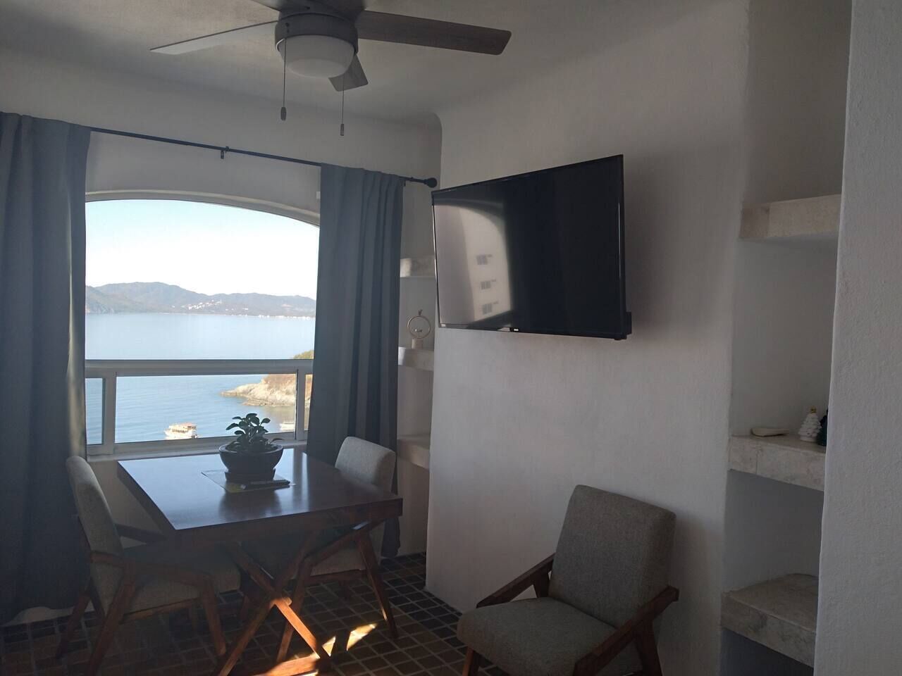 Ein Zimmer mit Esstisch, Stühlen, einem an der Wand befestigten Fernseher und Blick aufs Meer.