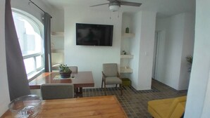 Ein Zimmer mit einem Holzesstisch, Stühlen, einem an der Wand befestigten Fernseher, einem Deckenventilator und einem Fenster mit Vorhängen.