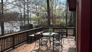 A wooden deck with a round table and chairs, surrounded by a railing, overlooking a body of water and trees.