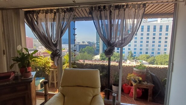 Ein Zimmer mit Blick auf eine Stadtlandschaft durch transparente Vorhänge, ein Sessel und Zimmerpflanzen.