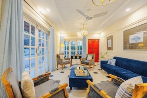 Un salon avec un canapé bleu, deux fauteuils, une table basse, une porte rouge et une grande fenêtre avec des rideaux.