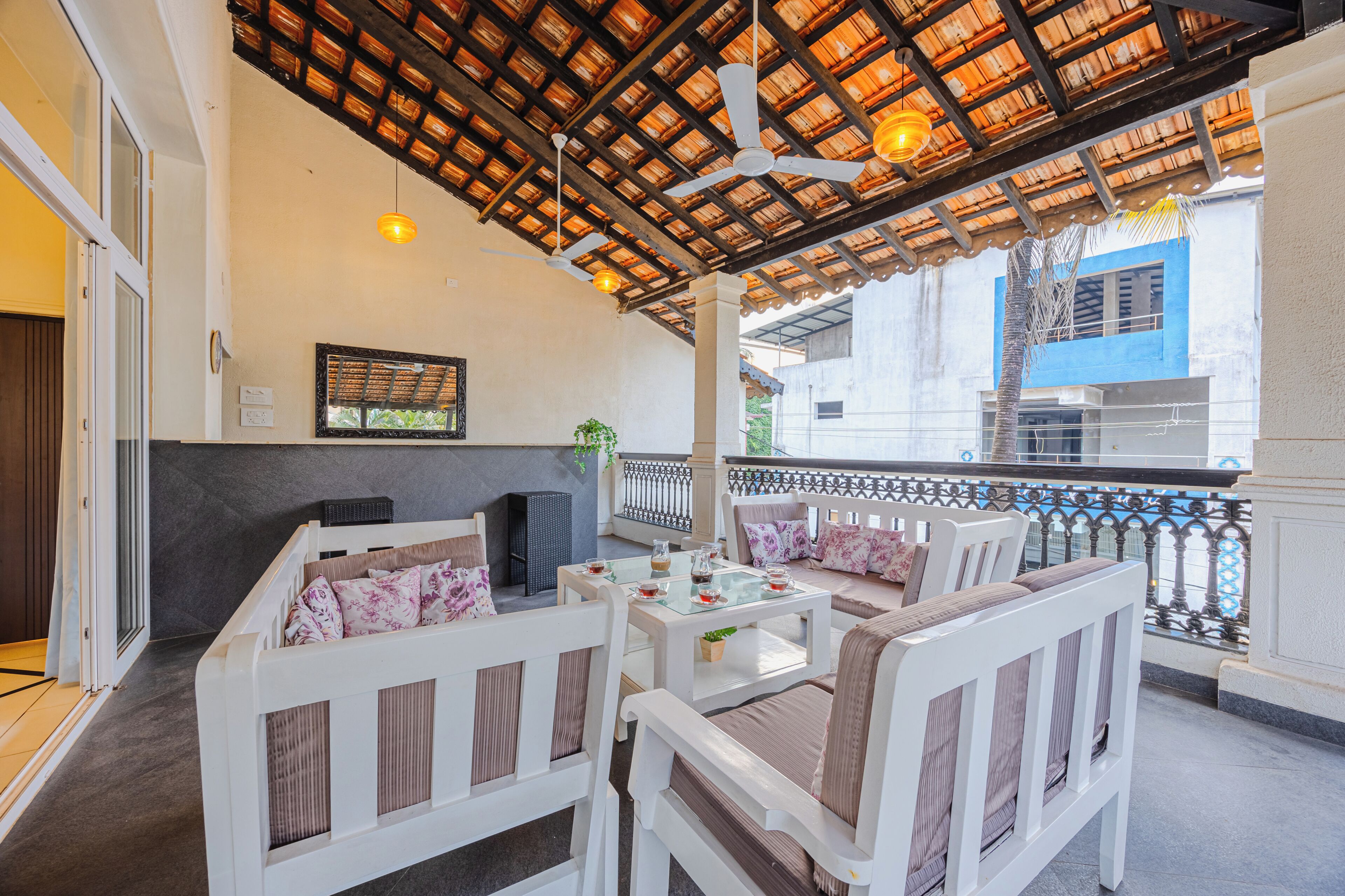 A covered outdoor seating area with white furniture, a ceiling fan, and a view of a building in the background.