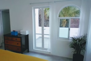 Ein Zimmer mit einer Schiebetür und einem Balkon, eine schwarze Kommode mit Schubladen, ein blauer Behälter und ein Blumentopf.