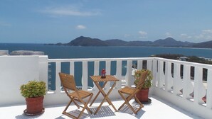 Un balcon avec deux chaises en bois et une petite table, offrant une vue sur la côte et les montagnes qui se dessinent à l’horizon.