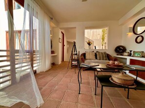 Un salón con sofá, una mesa de centro y una estantería. Hay una ventana con cortinas, un espejo en la pared y una puerta que da a otra habitación.