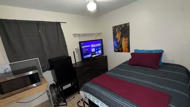 Une chambre à coucher avec un lit, une télévision, un bureau, un ventilateur de plafond et une affiche accrochée au mur.