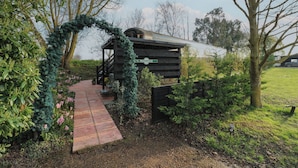 A wooden cabin with a covered porch, surrounded by lush greenery and a stone pathway leading to it. - The Carriage Ely Penzance (Ely)