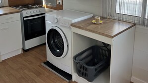 Een kleine keuken met een fornuis, oven, wasmachine en een tafeltje met een kopje.