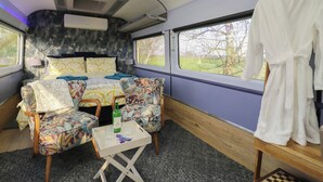 Ein Zugabteil mit einem Bett, zwei Stühlen, einem kleinen Tisch, einem Kleiderständer und einem Fenster mit Blick auf Bäume.