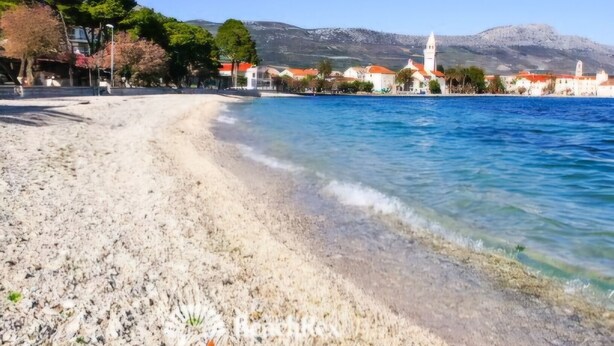 Una playa con agua cristalina, costa de guijarros y edificios al fondo.