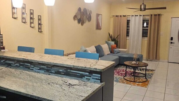 A modern kitchen with a central island, pendant lights, and a dining area with a sofa and a small round table. - Cozy 2-bedroom house where you can "Relax, Recharge, Repeat" (Bodden Town)