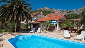 A poolside area with lounge chairs, a building with a red-tiled roof, and a mountainous backdrop. - Adriatic Guesthouse Balota with Shared Pool- Bungalow with Terrace (1) (Viganj)