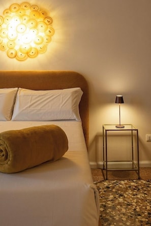 Una cama bien hecha con una toalla enrollada, un aplique de pared y una mesita con lámpara.