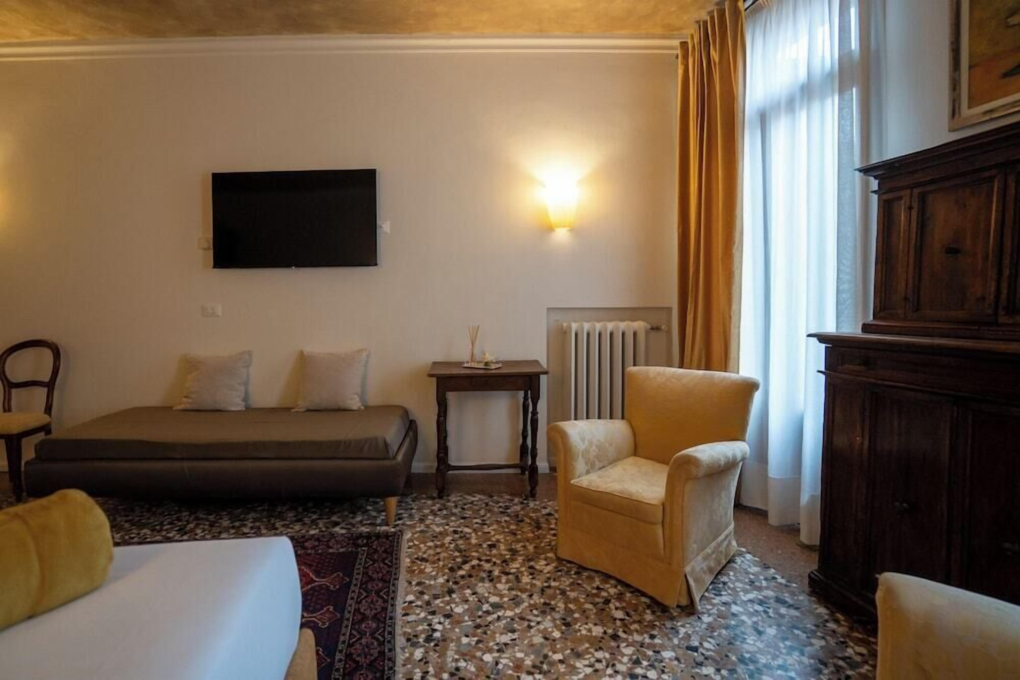 A hotel room with a bed, a sofa, a chair, a small table, a wall-mounted TV, and a radiator.