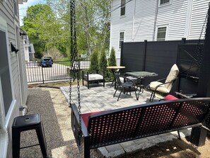 A patio with a swing, a table, and chairs, surrounded by a fence and a building.