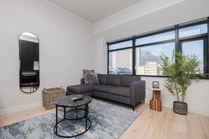 A modern living room with a gray sofa, a round coffee table, a large window with a city view, a mirror, and a potted plant. - 1205 at the Tokyo (Cape Town)