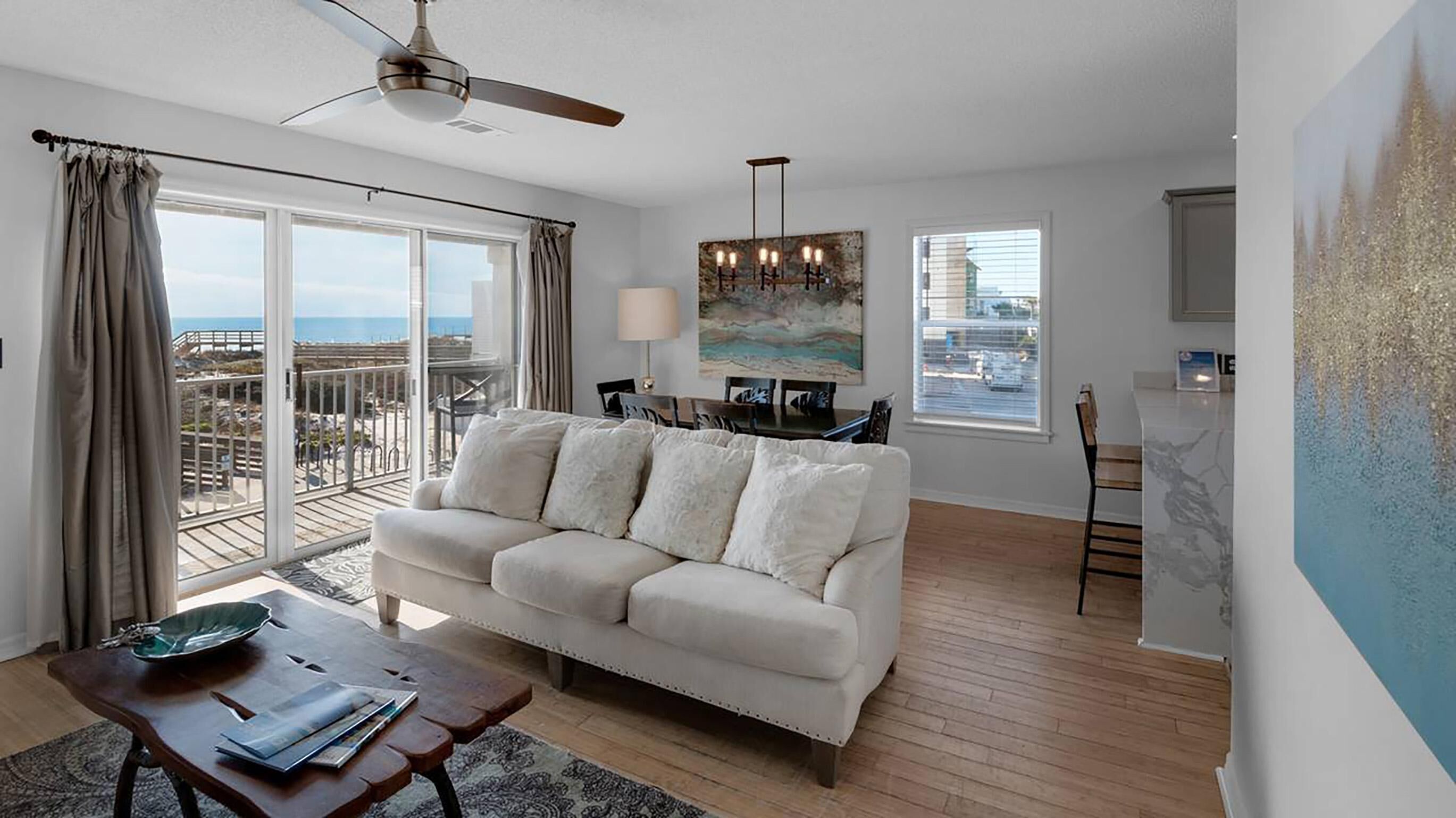 A living room with a sofa, a dining table, a ceiling fan, and a view of the ocean.