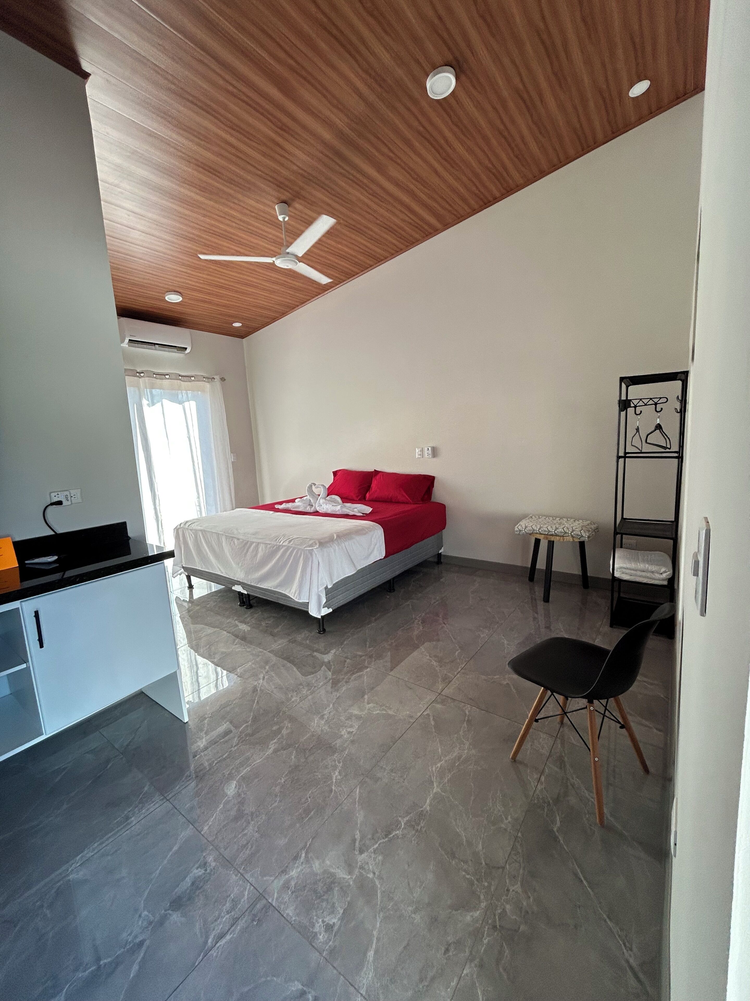 Een moderne slaapkamer met een bed, een plafondventilator, een klein krukje en een stoel.