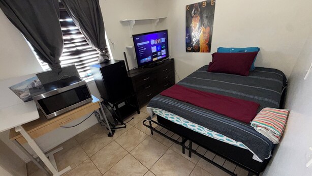 Ein Schlafzimmer mit einem Bett, einem Schreibtisch mit Computer, einem Fernseher und einem Poster an der Wand.