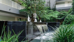Una fontana con acqua a cascata, circondata da vegetazione rigogliosa e un edificio a più piani con balconi.