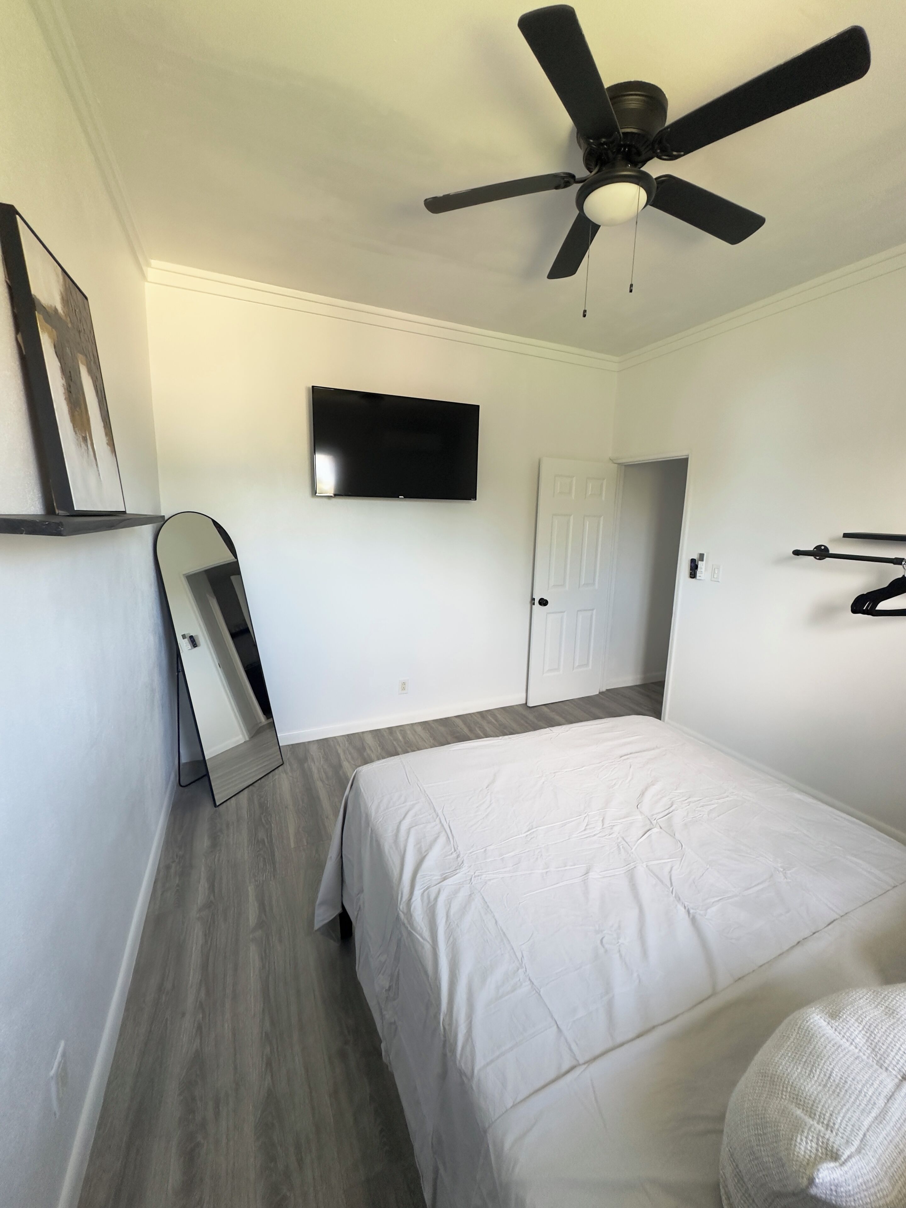 A bedroom with a bed, a ceiling fan, a TV, a door, and a mirror.