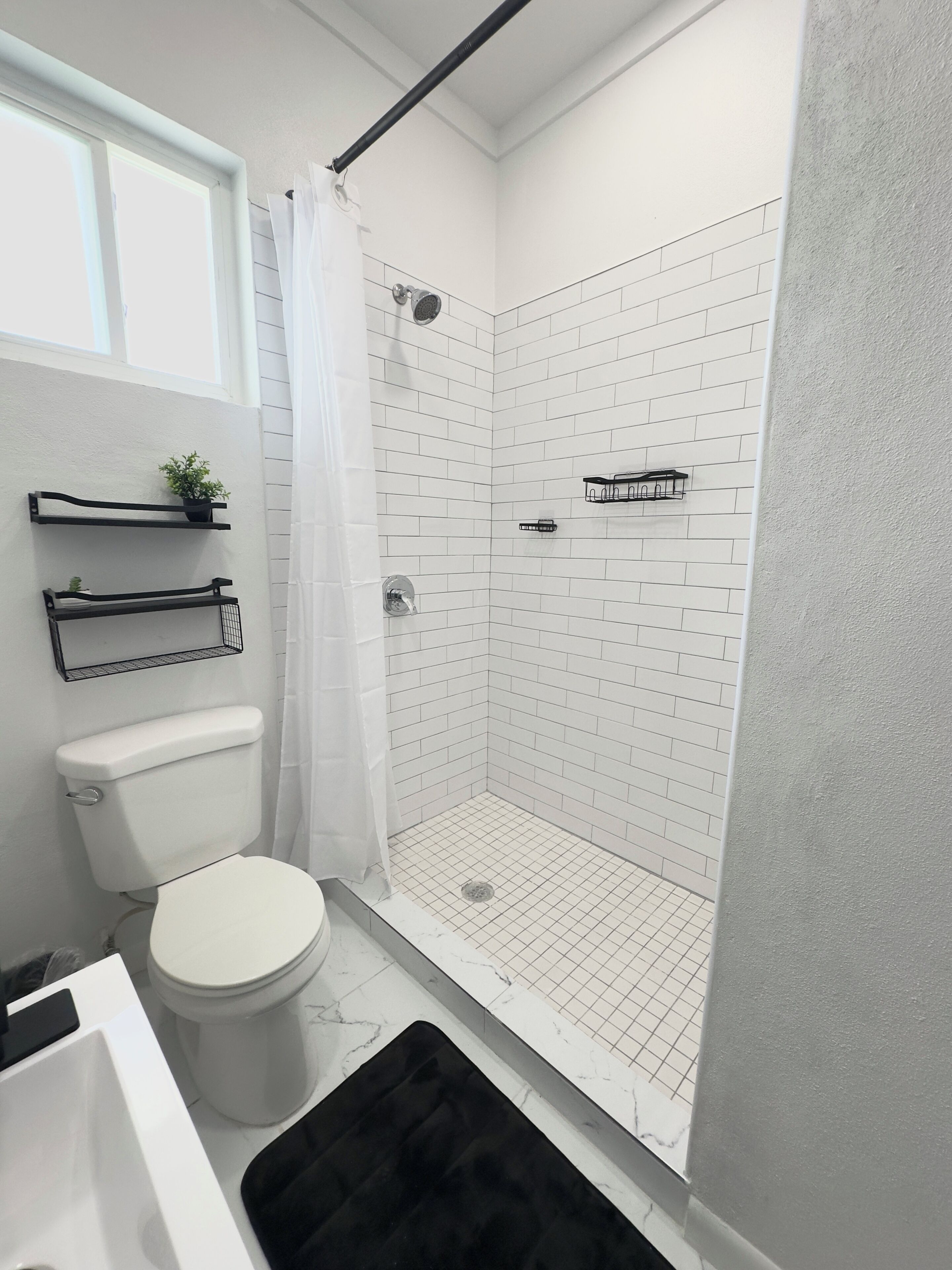 A bathroom with a white toilet, a shower with a white tiled wall, a black shower curtain, and a black rug.