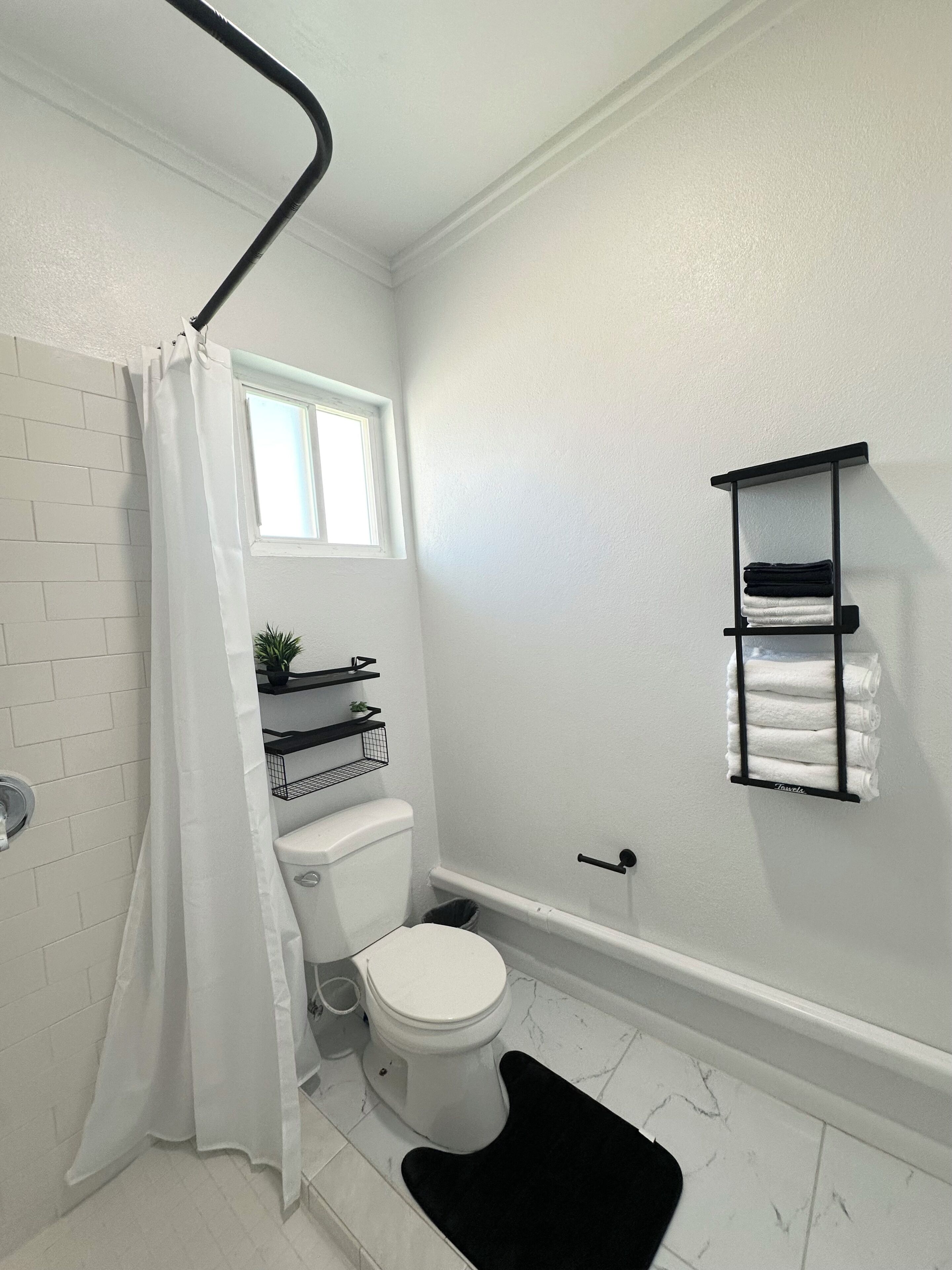 A bathroom with a white toilet, a black bath mat, a white shower curtain, a window, and a towel rack.