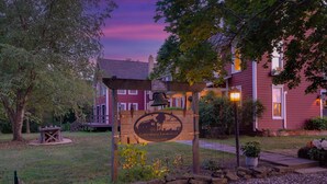 A wooden sign with a bell and a logo, a gravel path, and a red building with a porch. - The Farm Apartment at Gettysburg Farmstead (Mount Joy Township)