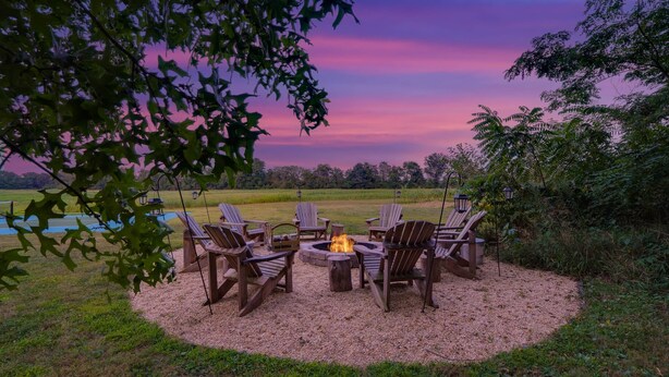 A circular seating area with chairs and a fire pit, surrounded by gravel, with a view of a body of water and trees in the background. - The Farm Apartment at Gettysburg Farmstead (Mount Joy Township)