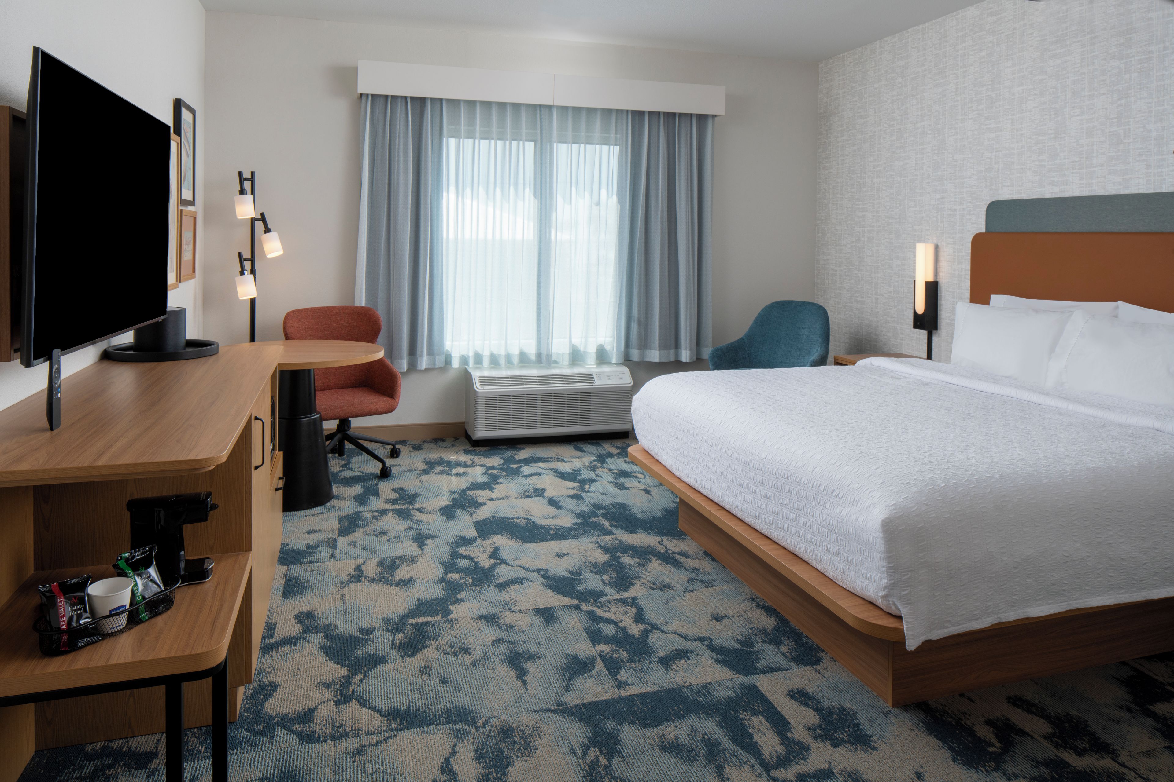 Habitación de hotel con cama, escritorio, silla, televisor y ventana con cortinas.