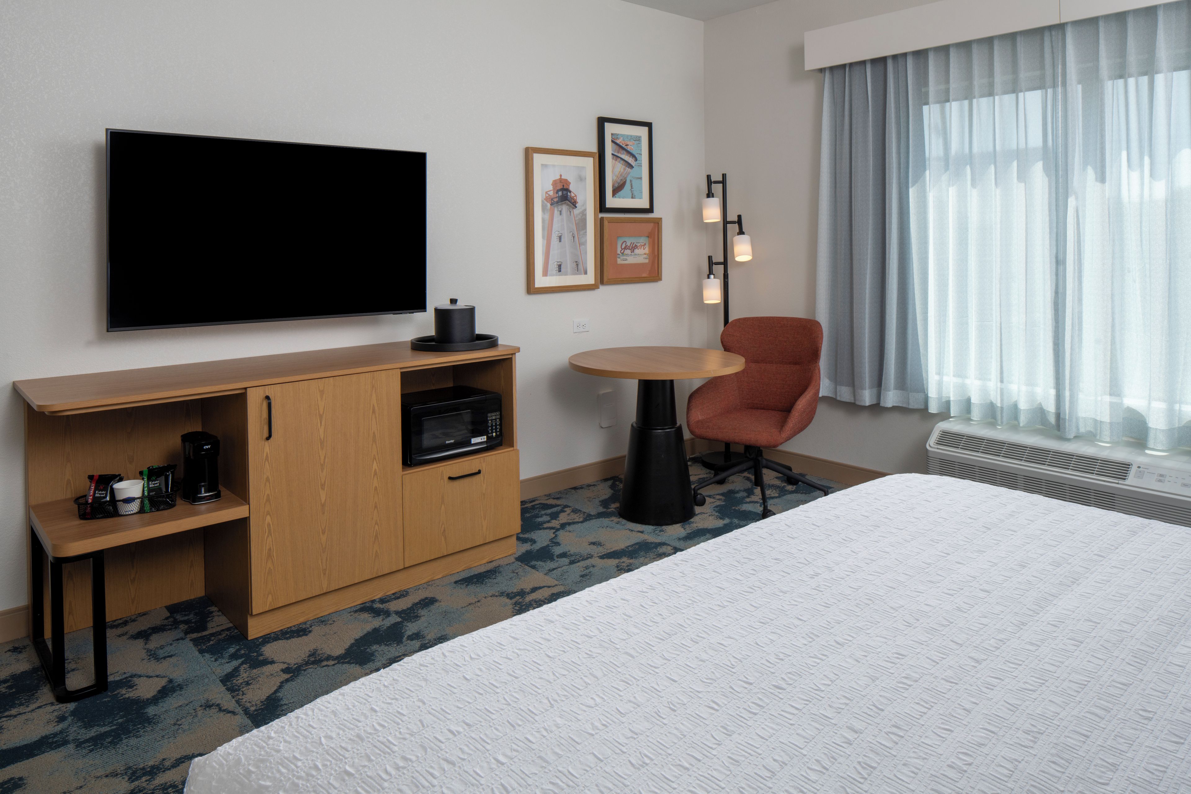 A hotel room with a bed, a TV, a desk with a microwave, a chair, and a window with curtains.