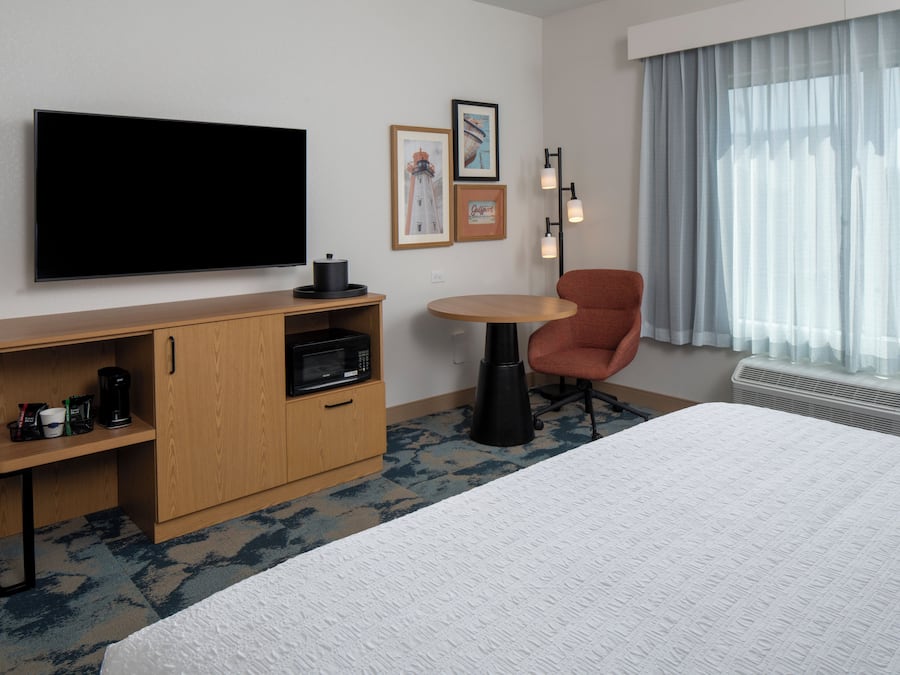 Ett hotellrum med en säng, en TV, ett skrivbord med en mikrovågsugn, en stol och ett fönster med gardiner.