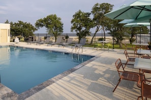 Une piscine entourée d’une clôture et d’arbres, avec des chaises longues et des tables à proximité.