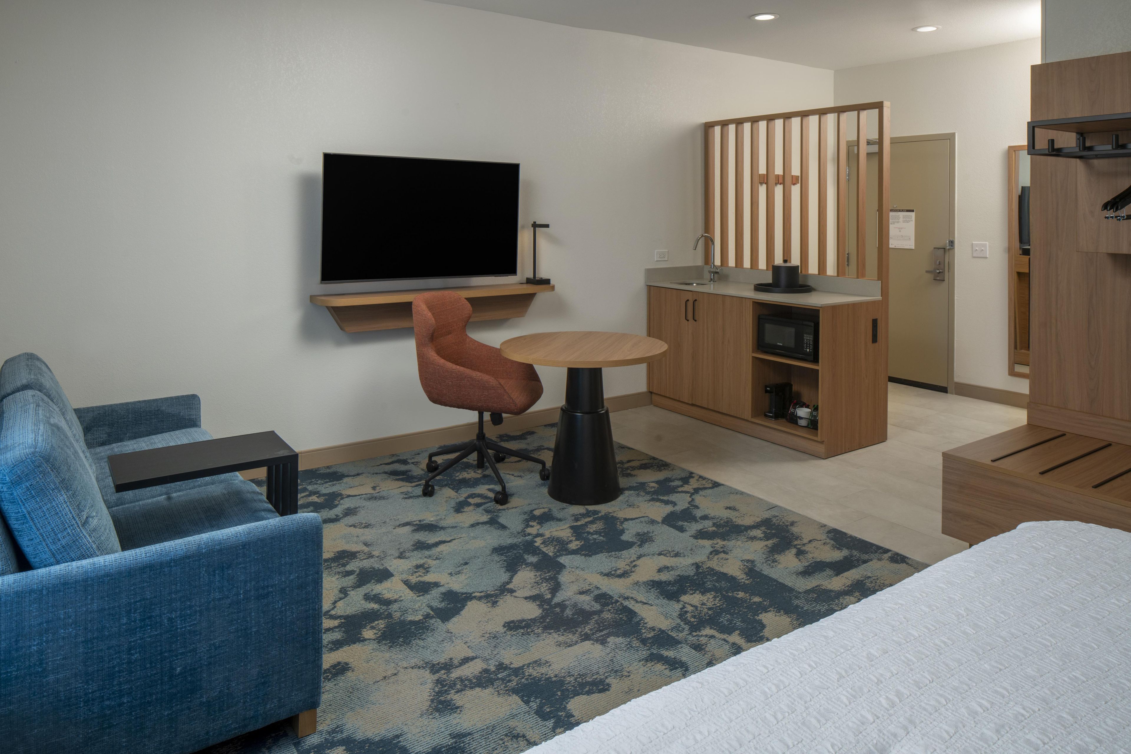 A hotel room with a bed, a blue armchair, a small round table, a television, a kitchenette, and a wardrobe.
