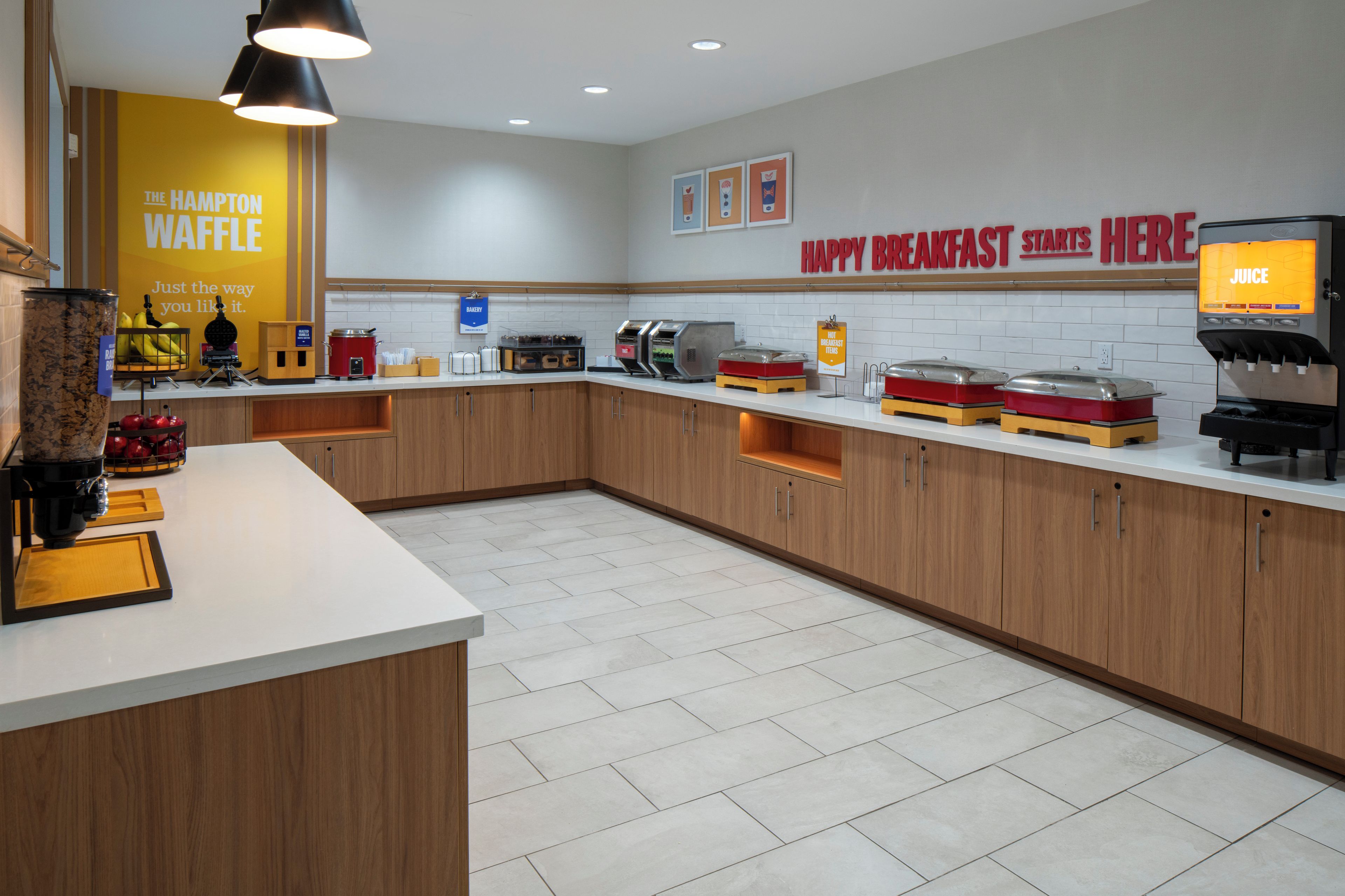 Un área de cocina moderna con una estación de waffles, un dispensador de jugo y un cartel que indica opciones para el desayuno.