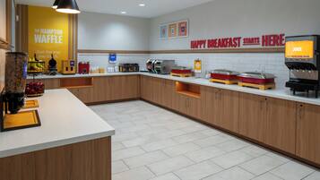 Un área de cocina moderna con una estación de waffles, un dispensador de jugo y un cartel que indica opciones para el desayuno.