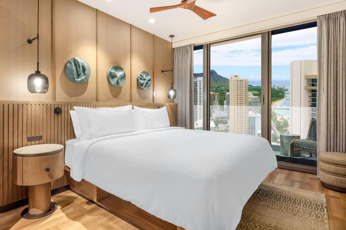 Hotelkamer met een groot bed, een houten hoofdeinde, een rond nachtkastje, een plafondventilator en uitzicht op een stadsgezicht door grote ramen.