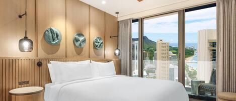 Una habitación de hotel con una cama grande, un cabecero de madera, una mesita de noche redonda, un ventilador de techo y vistas a un paisaje urbano a través de amplios ventanales.