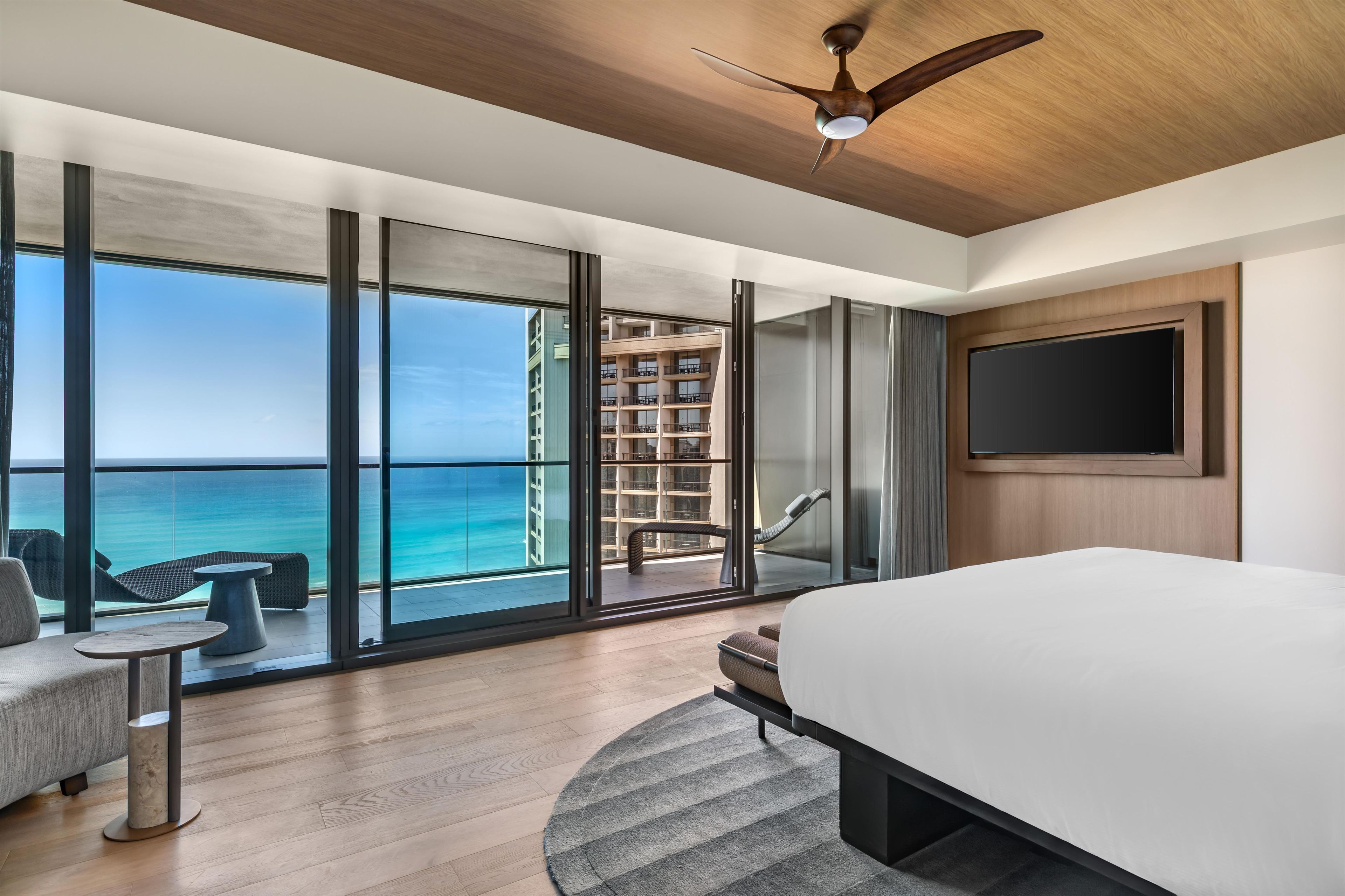 A modern hotel room with a large bed, a TV, and a balcony with a view of the ocean.