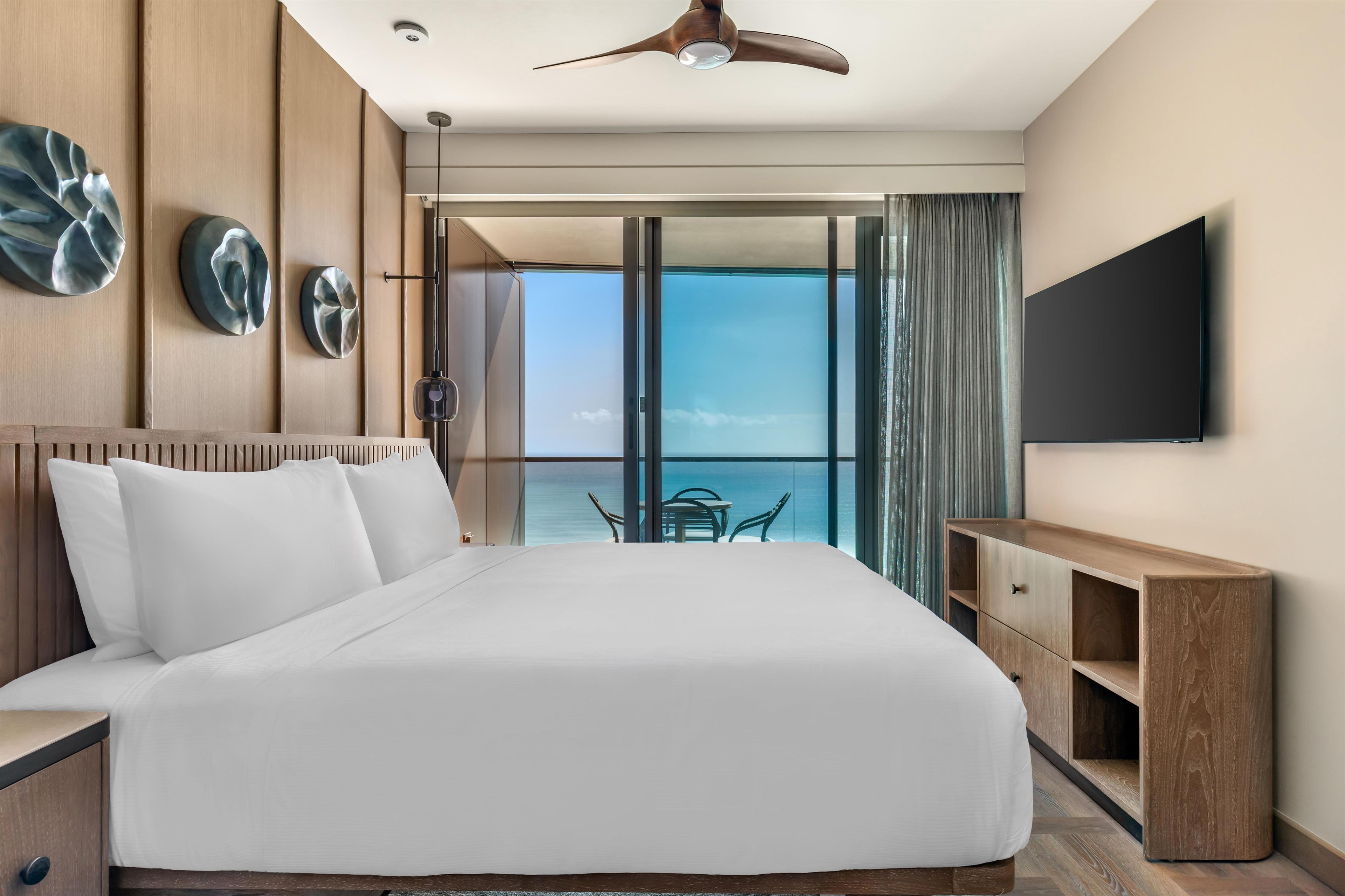 A hotel room with a large bed, a TV mounted on the wall, and a view of the ocean through the sliding glass doors.