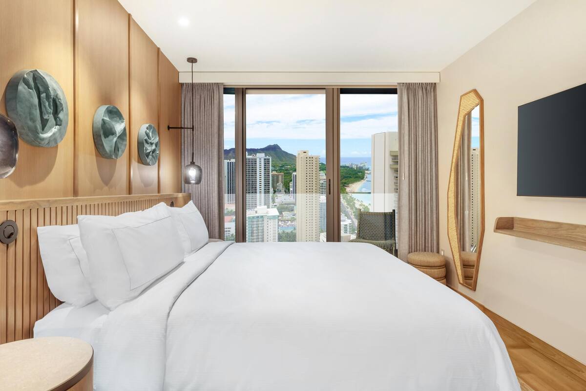 Een hotelkamer met een groot bed, houten lambrisering, uitzicht over de stad, een ronde tafel en een grote spiegel.