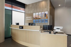 A modern hotel reception desk with a 'welcome to Hampton' sign, a digital display, and a glass door leading to the lobby. - Hampton Inn Long Island City  NY (Long Island City)