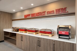 A breakfast station with a sign that reads 'HAPPY BREAKFAST STARTS HERE.' There are three covered food dispensers and a toaster oven on the counter. - Hampton Inn Long Island City  NY (Long Island City)