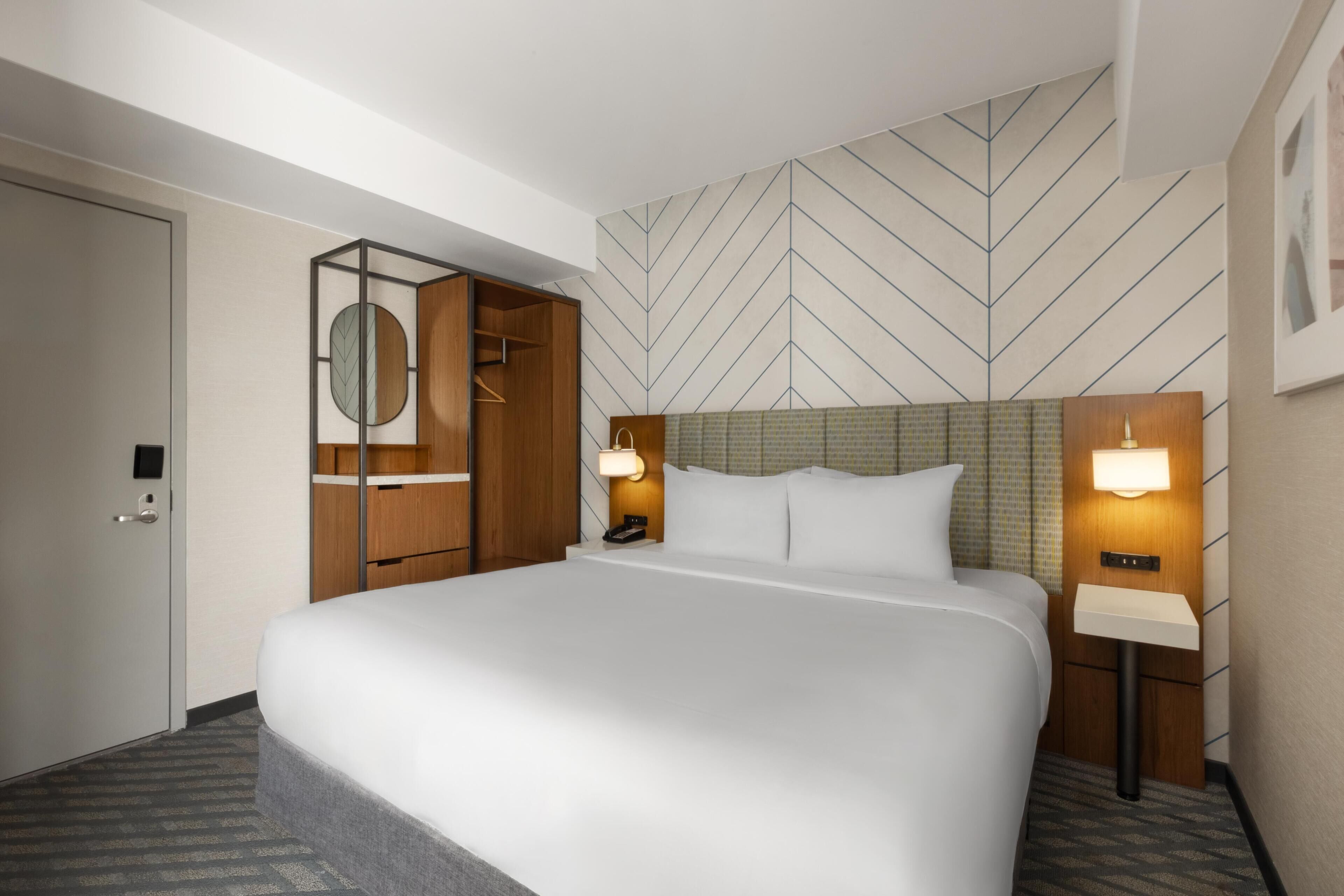 A hotel room with a large bed, a wardrobe, a mirror, bedside lamps, and a patterned wall.