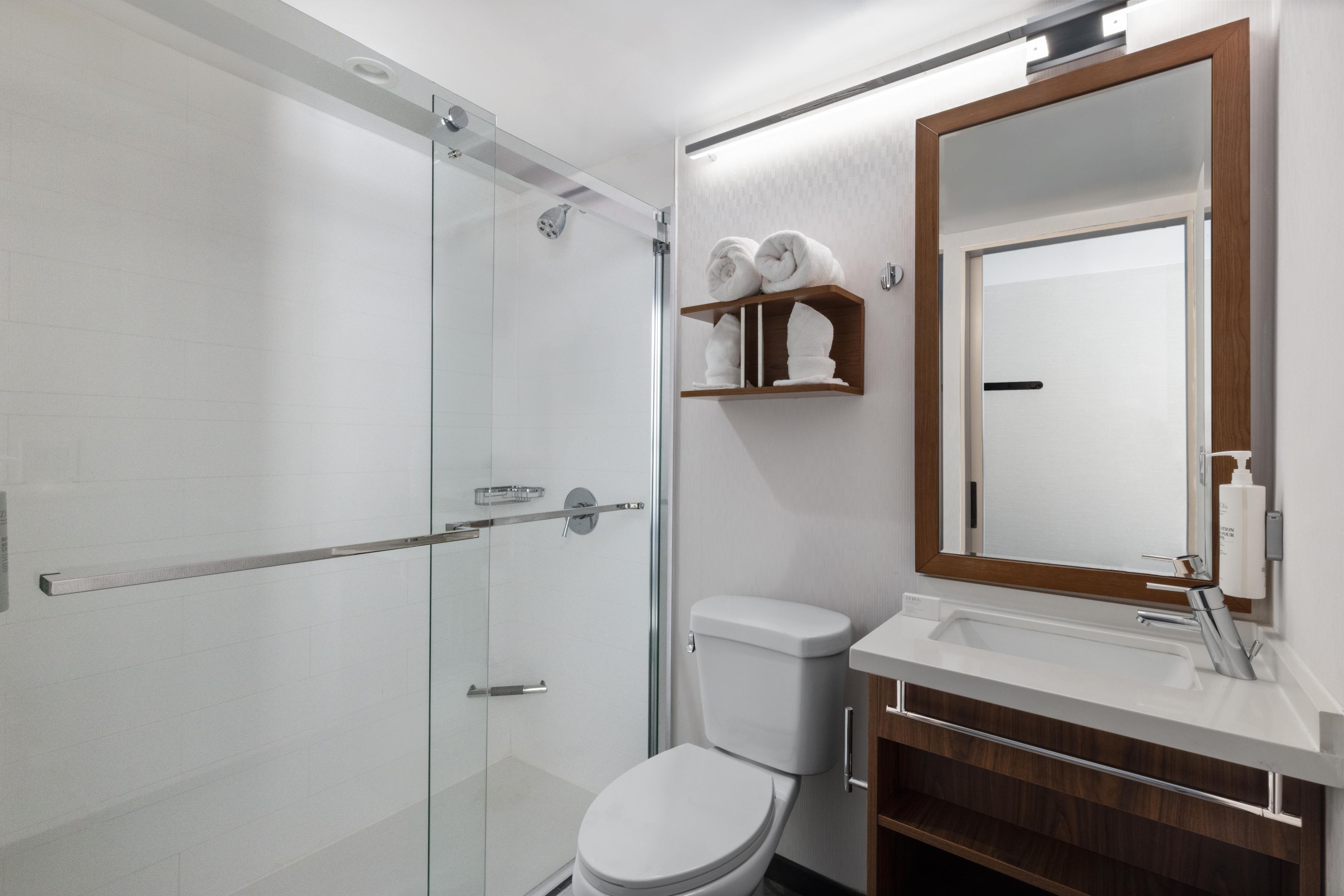 A modern bathroom with a glass shower enclosure, a white toilet, a wooden shelf with towels, a large mirror, and a white sink.