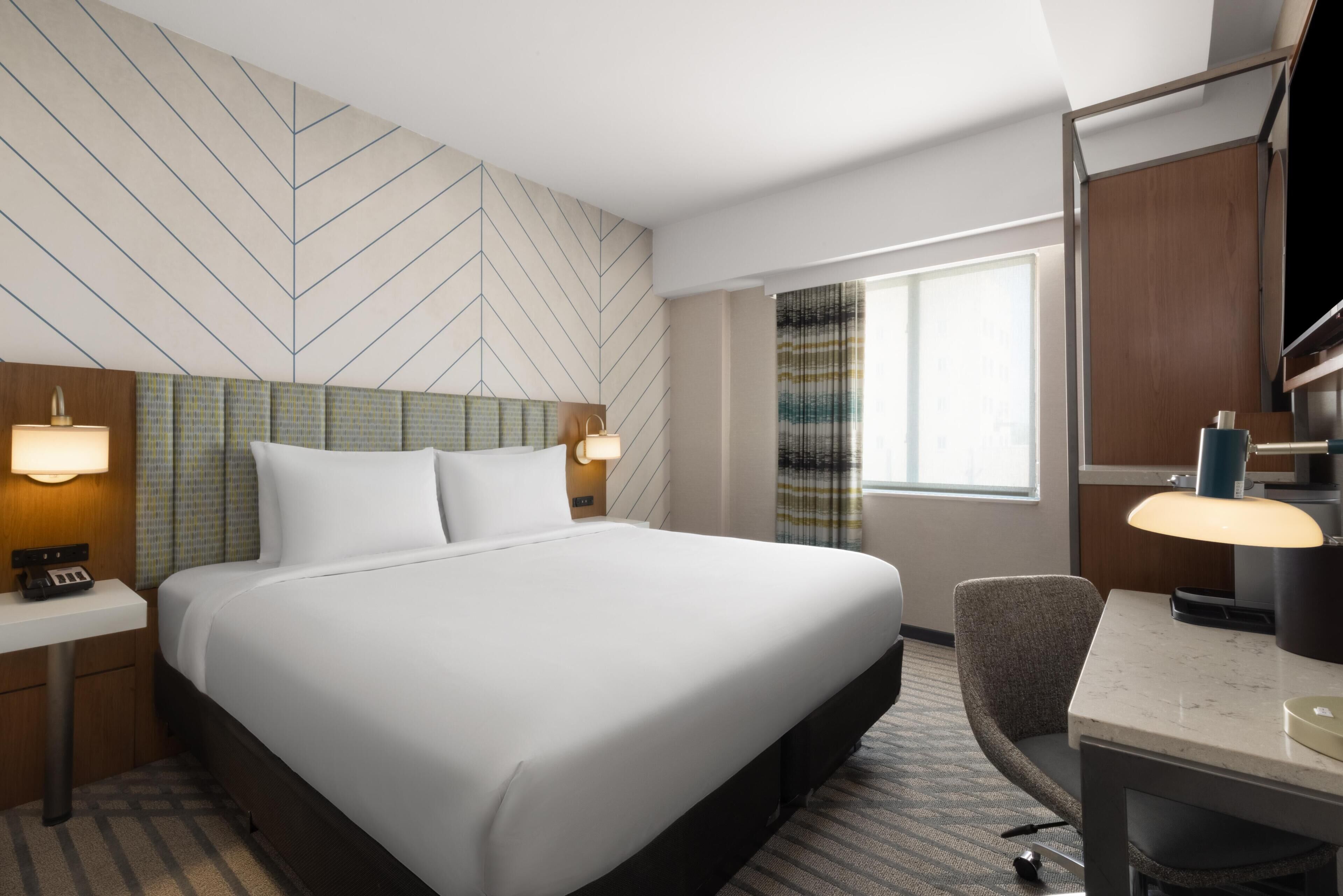 A hotel room with a large bed, a desk, a chair, a TV, and a window with curtains.