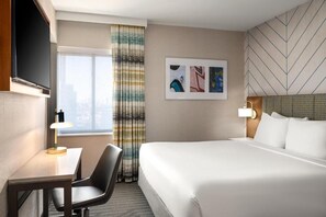 A hotel room with a bed, a desk, a chair, a TV, a window with curtains, and artwork on the wall. - Hampton Inn Long Island City  NY (Long Island City)