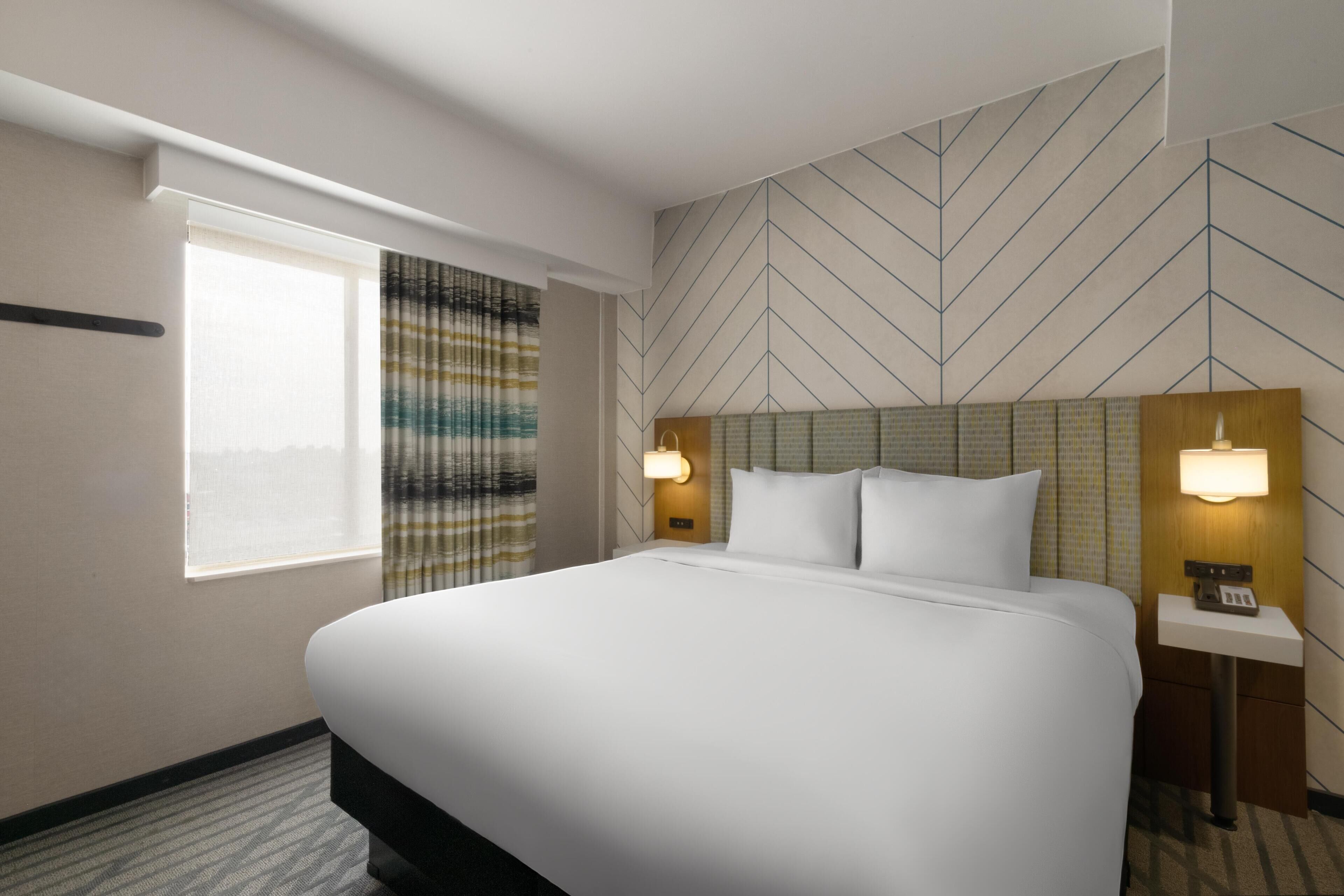 A hotel room with a large bed, two bedside lamps, a window with curtains, and a geometric wall pattern.